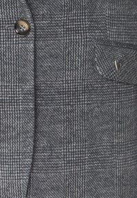 Gros plan d’un tissu à carreaux gris avec un bouton marron à gauche et une petite poche avec une épingle cœur argentée à droite.