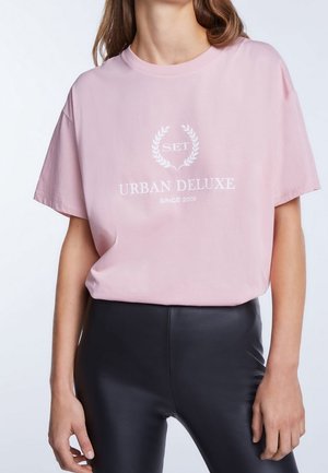 Lyserød bomulds T-shirt med en afslappet pasform, der har et hvidt logo og teksten "URBAN DELUXE" i blød, udekoreret skrifttype.