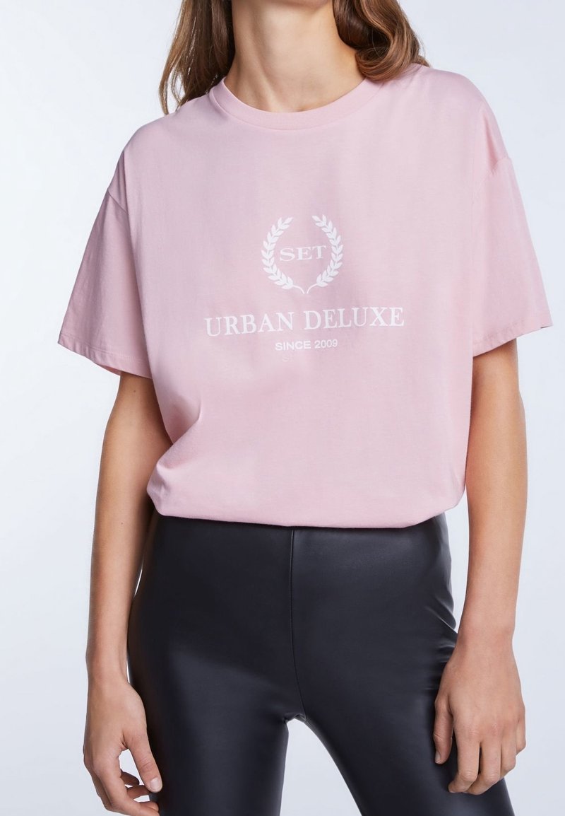 Světle růžové bavlněné tričko s volnějším střihem, s bílým logem a textem "URBAN DELUXE" v jemném, jednoduchém písmu.