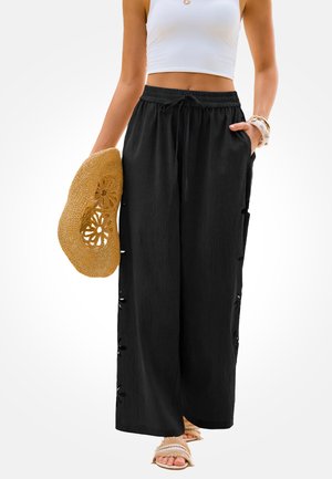 Frau trägt schwarze weit geschnittene Hose mit floralen Ausschnitten, weißes Croptop, hält einen geflochtenen Sonnenhut und beige Sandalen mit Fransen.