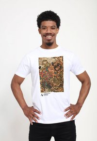 Hvid t-shirt med et farverigt grafisk print af en figur med tigre i en detaljeret, traditionel stil. Inkluderer museumsbranding i bunden.