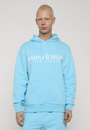 Person mit kurzen blonden Haaren, der einen hellblauen Hoodie und passende Hosen trägt, auf dem Hoodie steht "bruno banani".
