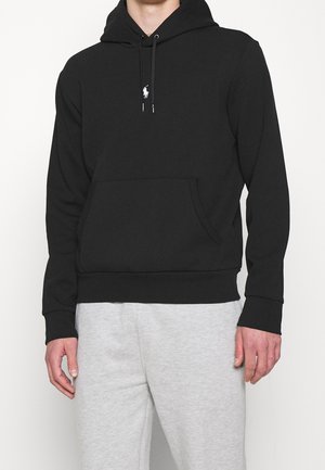 Man draagt een zwarte capuchontrui met voorvak en klein wit logo, gecombineerd met lichtgrijze trainingsbroek.