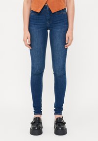 Kék, slim fit farmernadrág, magas derékkal, elülső zsebekkel és sötét varrással. Fekete, vastag talpú cipőkkel párosítva.