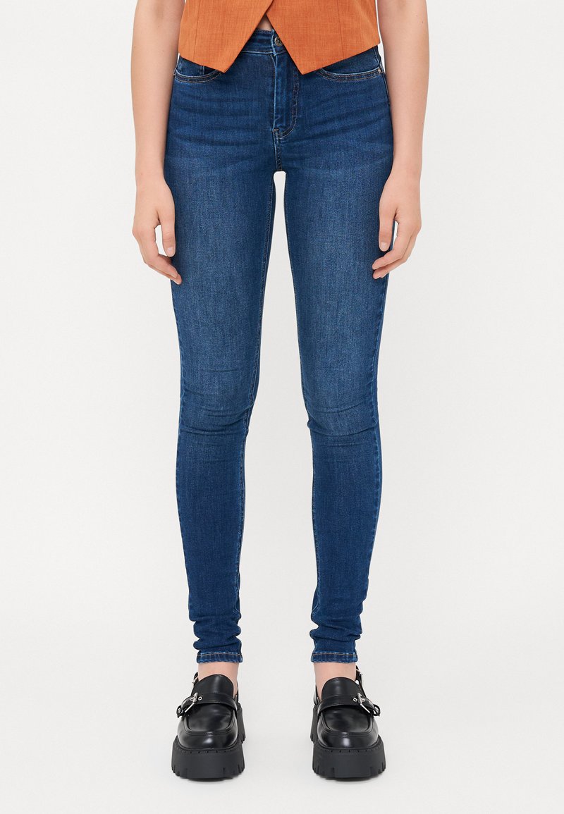 Kék, slim fit farmernadrág, magas derékkal, elülső zsebekkel és sötét varrással. Fekete, vastag talpú cipőkkel párosítva.