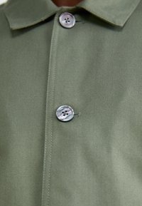 Nästa bild visar en närbild av olivgrön tyg, med en knapp med fyra hål och sömdetaljer, som framhäver en krage och en slät yta.