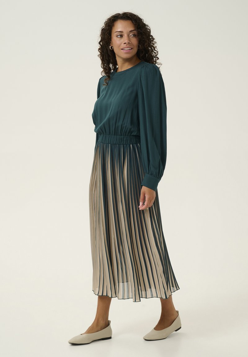 Larga vestido plisado con parte superior en color azul verdoso y falda de rayas beige; cuenta con una cintura ajustada y una tela ligera y fluida, combinado con zapatos planos beige.