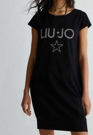 Kvinde iført en sort kjole med korte ærmer med "LIU JO" og et stjernedesign af rhinsten foran, stående mod en ensfarvet baggrund.
