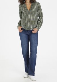 Grøn bluse med mønster, v-hals og lange ærmer, parret med blå vide jeans og hvide sneakers. Glat stof med et geometrisk design.
