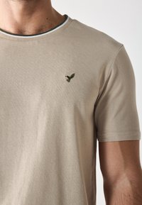 Bež majica od pamuka s kratkim rukavima, koja ima zeleni izvezeni logo ptice na lijevoj strani prsiju i rebrasti ovratnik s kontrastnim prugama.