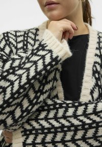 Cardigan tricoté en beige et noir avec un motif géométrique, doté d'un col en V et de bords côtelés. Matière douce en mélange de laine texturée.