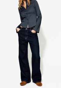 Marineblå skjorta med knapp-up och långa ärmar, kombinerad med vida mörka jeans med kontrasterande vit sömnad och klassisk knappstängning.