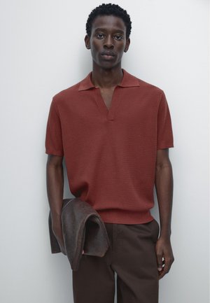 Homme portant un polo en maille à manches courtes couleur rouille et un pantalon marron foncé, tenant une veste en cuir marron pliée devant un fond uni clair.