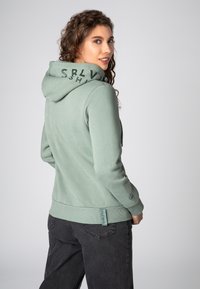 Lichtgroene hoodie met een textuurafwerking, voorzien van een trekkoordkap en een logo op de rug. Gecombineerd met donkergrijze broeken die een getailleerd ontwerp laten zien.