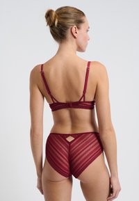 Vrouw met blond haar in een knot, gekleed in een bordeauxrode kanten bh en bijpassende hoge doorschijnende kanten slip, met de rug naar de camera gekeerd tegen een witte achtergrond.