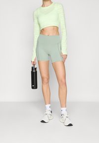 Light green cropped long-sleeve top with textured detailing, paired with grey shorts. Holding a black water bottle, wearing white sneakers and socks.