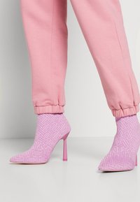 Botas de tacón aguja rosa hasta el tobillo con un patrón texturizado geométrico, puntera afilada y un tacón rosa a juego, combinadas con pantalones de chándal rosa con puños.