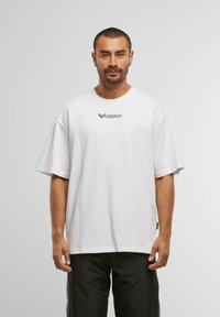 Mann med kort hår og skjegg iført en hvit t-skjorte med "Colucci"-logo og svarte bukser, stående mot en enkel lys bakgrunn.
