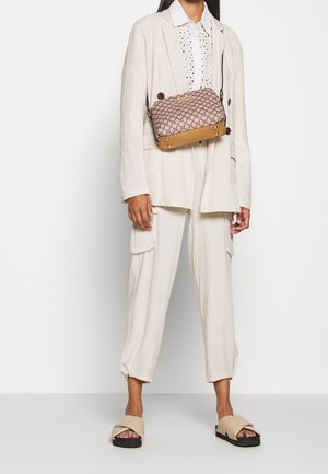 Persona che indossa un completo in lino beige con pantaloni cargo, una camicia bianca con fori a bottone e una borsa a tracolla a fantasia color tan, oltre a sandali beige con suola nera.