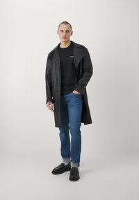 Schwarzer Leder-Trenchcoat über einem schwarzen T-Shirt, kombiniert mit blauen Jeans und schwarzen Slippern. Das Modell steht vor einem schlichten Hintergrund.