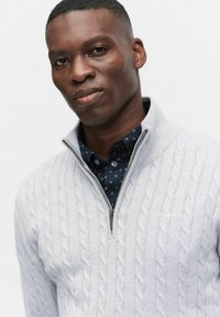 Jasnoszary sweter pleciony z półsuwnym kołnierzem założony na ciemną, wzorzystą koszulę. Sweter ma teksturowane detale oraz subtelne logo na piersi.