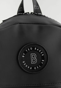Crna torba sa glatkom teksturom, s okruglom oznakom koja sadrži slovo "B" i tekst "BY TED BAKER". Uključuje zatvaranje na patentni zatvarač i ručku na vrhu.