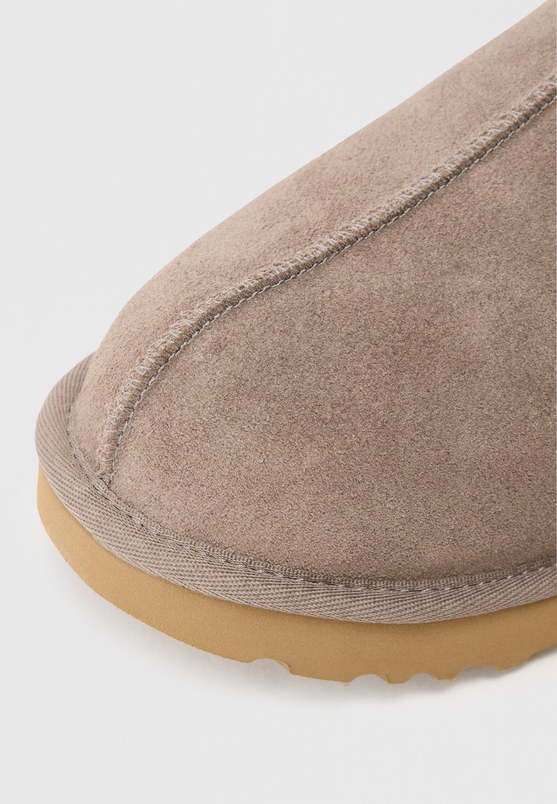 Acercamiento de la punta de un zapato de ante beige con costuras visibles y suela de goma marrón claro sobre un fondo blanco.