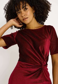 Haut à manches courtes bordeaux en tissu texturé, avec un détail de nœud torsadé à la taille. Fini lisse avec un éclat subtil.