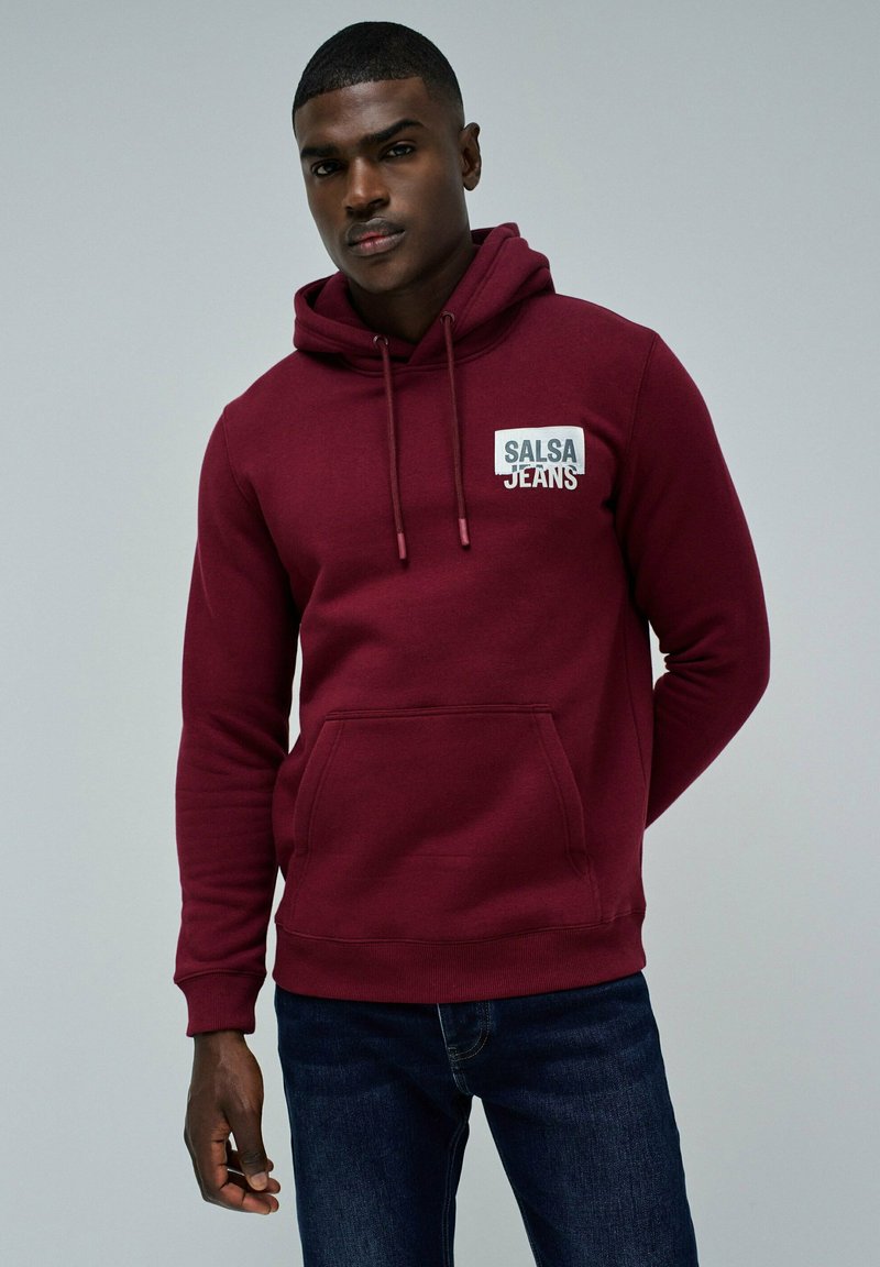 Młody mężczyzna ubrany w bordowy hoodie z napisem "SALSA JEANS" na klatce piersiowej, w połączeniu z ciemnoniebieskimi dżinsami, stoi na tle jednolitym.
