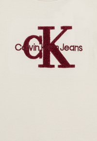 Camiseta color crema con un gran logo bordado en burdeos "CK" y el texto "Calvin Klein Jeans" debajo en una fuente moderna.