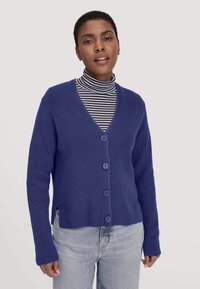 Frau in einem blauen Knopfkarteigan über einem schwarz-weiß gestreiften Rollkragenpullover und hellblauen Jeans, die vor einem einfarbigen Hintergrund steht.