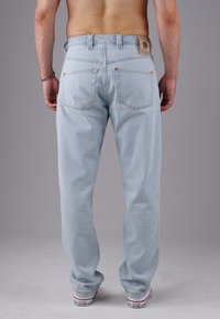 Mann in hellblauen Jeans und weißen Converse-Sneakern, der gegen einen schlichten grauen Hintergrund steht, von hinten gezeigt.