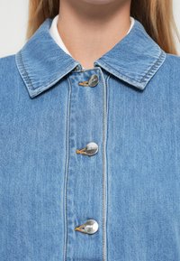 Nærbilde av lyseblå denimjakke med sølvknapper og stor krage, båret over en hvit skjorte.