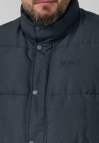 Man die een donkerblauwe gewatteerde jas met hoge kraag en drukknoopsluiting draagt, met een patch "Elevated Casual Est 1969" op de borst.