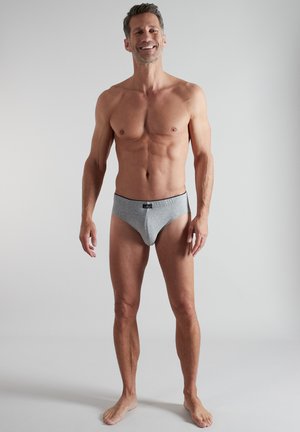 Lächelnder Mann, der barfuß steht und eng anliegende graue Unterhosen vor einem einfachen hellen Hintergrund trägt.