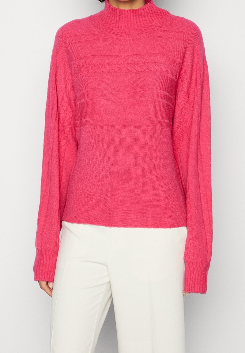 Femme portant un pull en maille torsadée rose vif avec un col et des poignets côtelés, associé à un pantalon blanc cassé sur un fond uni.