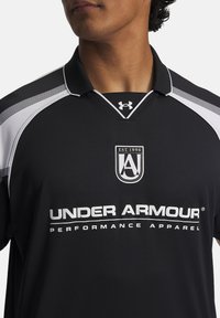 Polo de manga corta negro con acentos blancos y cuello tipo polo. Presenta el logo de Under Armour y la marca de ropa de rendimiento.