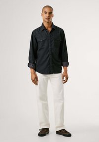 Dunkelblaues Denim-Hemd mit zwei Brusttaschen, Knopfleiste und hochgekrempelten Ärmeln; kombiniert mit weißen Jeans und dunklen Schnürschuhen.