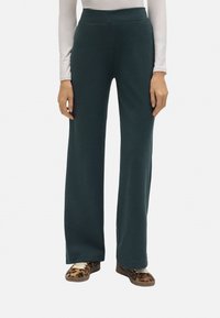 Pantalones de pierna ancha verde hechos de un material suave con una cintura alta. Combinados con una blusa blanca de manga larga y calzado de estampado animal.