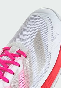 Białe sportowe buty z siateczkową cholewką, różowymi sznurówkami i fakturowanym wzorem oraz wyraźną czerwoną gumową podeszwą. Zauważalne odblaskowe akcenty.