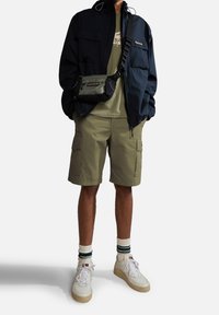 Veste coupe-vent marine avec plusieurs poches, shorts cargo olive, baskets blanches et sac bandoulière. Tissu texturé avec des couleurs contrastées.