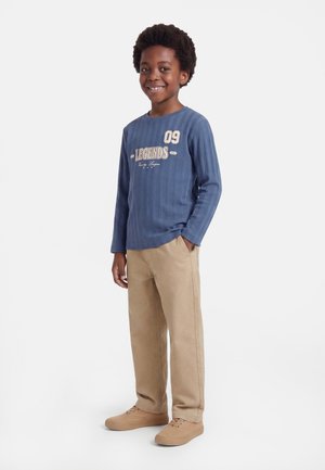 Lächelnder Junge steht mit den Händen in den Taschen, trägt ein blaues Langarmshirt mit Text sowie beige Hose und Schuhe vor weißem Hintergrund.