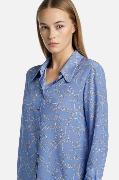Lichtblauwe shirt met een zijden textuur en witte geborduurde tekst in een cursieve patroon. Met knopen en een klassiek kraagontwerp.