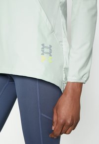 Chemise longue vert clair à manches longues avec un ourlet arrondi et un logo Under Armour en gris et jaune. Portée sur des leggings texturés bleu marine.