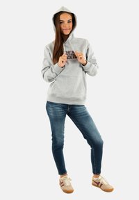 Felpa grigia chiara con tasca a marsupio e logo scuro, abbinata a jeans blu e sneakers beige con righe marroni.