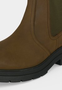 Bottine en cuir marron avec semelle en caoutchouc noire et panneau latéral élastique, montrant des coutures détaillées et une semelle texturée.