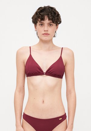 Frau mit kurzen lockigen Haaren, die ein burgunderfarbenes, geripptes Bikini-Oberteil und passende Unterteile vor einfachem weißem Hintergrund trägt.