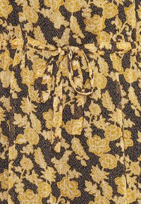 Tissu noir avec motif floral jaune et blanc présentant des feuilles et des fleurs détaillées, noué au centre avec un cordon assorti en nœud.