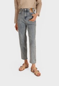 Jeans azzurri chiari a vita alta con gamba dritta, abbinati a sandali aperti beige con accento a fibbia e straps intrecciati. Maglione beige lavorato a maglia.