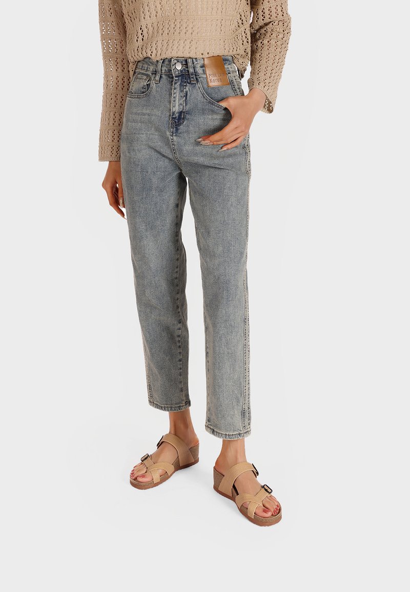 Jeans azzurri chiari a vita alta con gamba dritta, abbinati a sandali aperti beige con accento a fibbia e straps intrecciati. Maglione beige lavorato a maglia.